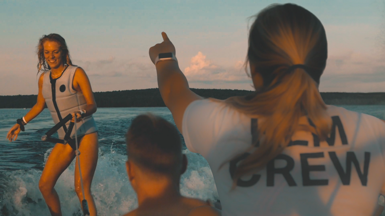 Person waterskiing with another person pointing towards the water at sunset.