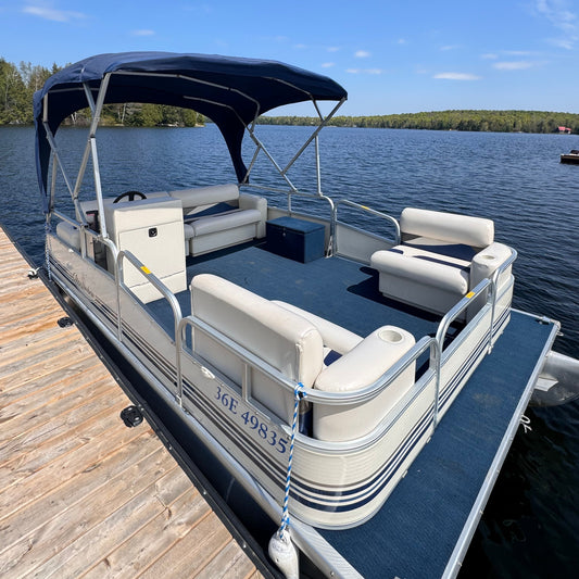 A Misty Harbour pontoon boat docked at a wooden dock, with a canopy top and seating arrangement visible, against a backdrop of a lake and trees.